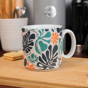 Puede incluir: Taza de cerámica blanca con un colorido diseño floral en verde azulado, naranja y azul marino. La taza presenta texto inspirador y un asa blanca. La taza está sobre una tabla de cortar de madera.