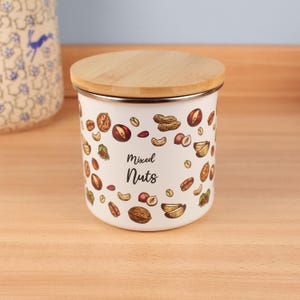 Op de afbeelding: Een witte emaille voorraadpot met een bamboe deksel, versierd met illustraties van gemengde noten. De pot heeft de tekst "Mixed Nuts" erop. Geschikt voor het bewaren van droge waren in de keuken.