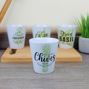 Puede incluir: Maceteros de cerámica blanca con texto e ilustraciones verdes. Una maceta dice "Chives" con una ilustración verde de cebollino. Otras macetas dicen "Coriander", "Hot Chili" y "Green Basil".