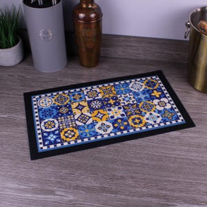 Peut inclure: Un tapis de cuisine rectangulaire présentant un motif mosaïque vibrant de carreaux bleus, jaunes et blancs. Le design se compose de motifs géométriques et floraux répétés disposés en grille, encadrés par une bordure noire unie.
