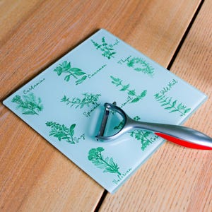 Puede incluir: Una tabla de cortar de vidrio azul claro con una variedad de hierbas y especias impresas en ella. La tabla está decorada con ilustraciones de albahaca, tomillo, canela, berro, espinacas, salvia, menta, menta silvestre, cilantro y valeriana. Un pelador de verduras plateado y rojo está sobre la tabla.