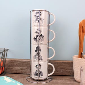 Può includere: Una pila di quattro tazze in ceramica bianca con ritratti in bianco e nero di comici classici, tenute in un supporto in metallo argentato. Le tazze hanno manici bianchi e sono impilate verticalmente. Sono visibili anche utensili da cucina e un cestino di filo metallico con oggetti arancioni.
