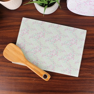 Peut inclure: Une spatule en bambou est posée sur une planche à découper rectangulaire en verre. La planche a un fond vert clair avec un motif floral rose. La spatule est marron clair avec le mot "TYPHOON" imprimé sur le manche.