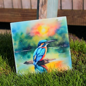 May include: A watercolour painting of a kingfisher perched on a rock in a lake. The bird is blue, green, and orange with a long, pointed beak. The background is a sunset with orange, pink, and purple hues.
