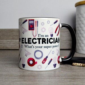 May include: White ceramic mug with a black rim and handle. The mug features a design of electrical tools and the text "I'm an Electrician. What's your super power?"