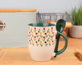 May include: A white ceramic mug with a green interior and a matching spoon. The mug is decorated with a pattern of colorful tulips. The spoon handle curves around the mug, creating a unique design. The mug sits on a wooden cutting board.