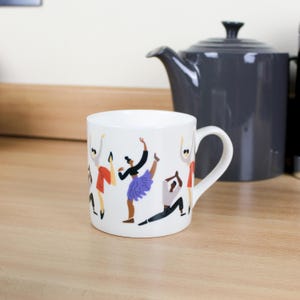 May include: White ceramic mug featuring a colourful illustration of dancers in various poses. The mug has a handle on the right side.