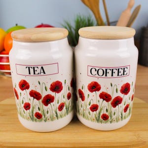 Op de afbeelding: Twee witte keramische potten met houten deksels, met de labels "TEA" en "COFFEE". Elke pot heeft een bloemenontwerp van rode klaprozen en groene stelen. De potten staan op een houten oppervlak.