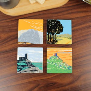 Puede incluir: Juego de cuatro posavasos cuadrados con diseños de paisajes. Cada posavasos presenta una escena diferente: una montaña, una hilera de árboles, un castillo costero y una cima montañosa. Los posavasos tienen un acabado brillante y están sobre una superficie de madera.