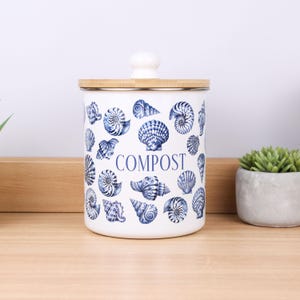 May include: A white cylindrical compost bin featuring a repeating pattern of blue seashells and the word COMPOST printed in blue. The container has a flat wooden lid with a small white knob handle centred on top of the lid.