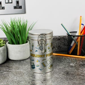 Peut inclure: Une boîte métallique argentée avec un couvercle à charnière et un fermoir. La boîte est décorée d'illustrations bleues et jaunes et des mots "HOLIDAY FUN". La boîte est sur un comptoir gris.