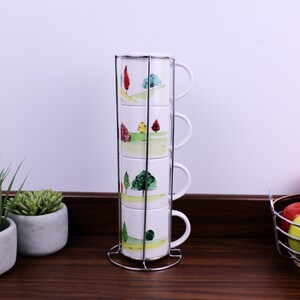 Puede incluir: Un juego de cuatro tazas de cerámica blanca apiladas verticalmente en un soporte de alambre de metal cromado. Cada taza presenta un estampado de paisaje estilo acuarela con colinas verdes y árboles en tonos rojos, amarillos y verdes.