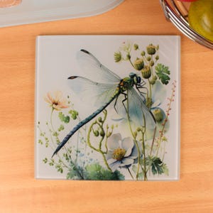Puede incluir: Posavasos cuadrado de vidrio con una ilustración de acuarela de una libélula con alas translúcidas, entre flores blancas y melocotón y follaje verde. El posavasos tiene un acabado brillante.
