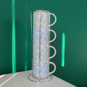 Puede incluir: Juego de cuatro tazas apiladas de cerámica blanca con un diseño de hojas azules y verdes, sujetas en un soporte de metal cromado. Las tazas tienen asas blancas y están sobre un fondo verde.