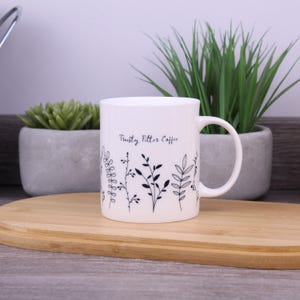 Può includere: Tazza in ceramica bianca con un design botanico nero e la scritta "Trusty Filter Coffee". La tazza ha un manico ed è appoggiata su un tagliere di legno. Piante decorative sullo sfondo.