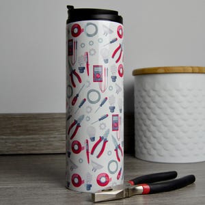 May include: White travel mug with a black lid and a pattern of red and blue electrician's tools. The mug is sitting on a wooden surface.