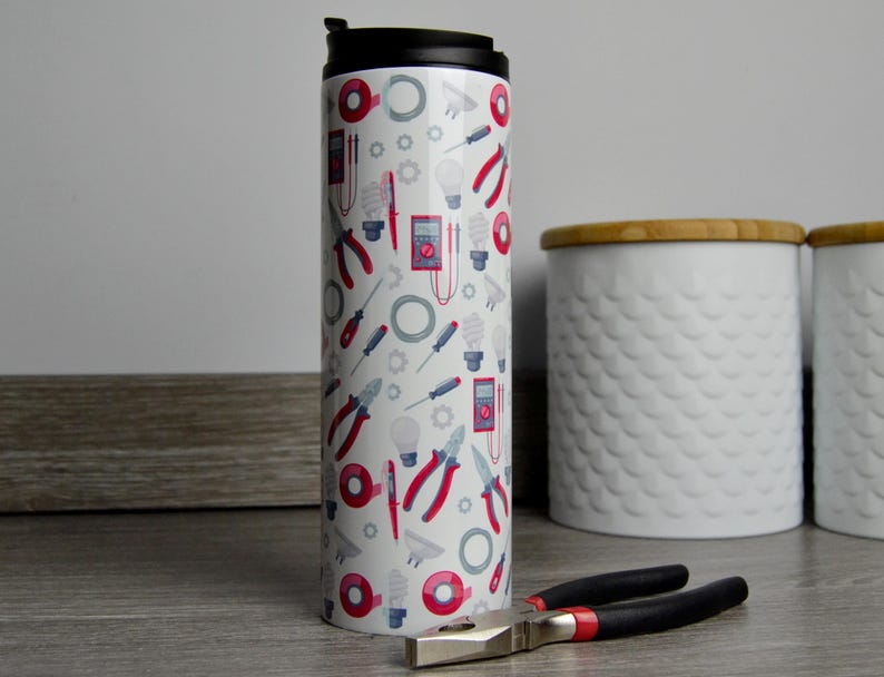 May include: White travel mug with a black lid and a pattern of red and blue electrician's tools. The mug is sitting on a wooden surface.