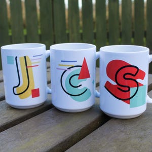 Può includere: Tre tazze in ceramica bianca con disegni geometrici in giallo, rosso, blu e verde. Le tazze compongono la parola "JCS" in uno stile retrò.