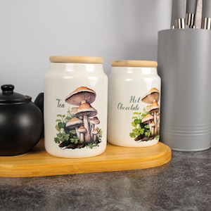 Può includere: Due barattoli in ceramica color crema con coperchi in legno, decorati con illustrazioni di funghi. Uno recita "Tea" e l'altro "Hot Chocolate". Sono su un vassoio di bambù, accanto a una teiera nera e un portautensili grigio.