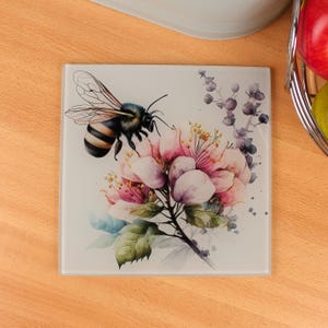 Peut inclure: Sous-verre carré en verre avec un motif aquarelle d'une abeille et de fleurs roses. L'abeille a des rayures noires et jaunes et est positionnée près des fleurs. Les fleurs ont des pétales roses et blancs avec des feuilles vertes et des accents violets.