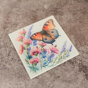 Può includere: Sottobicchiere quadrato in vetro con una farfalla colorata con ali arancioni, blu e marroni. Il sottobicchiere presenta un motivo floreale con fiori rosa e blu. Lo sfondo è bianco.