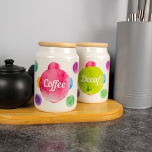 Può includere: Due barattoli di caffè in ceramica bianca con coperchi in legno. Un barattolo ha un design ad acquerello rosa con la scritta "Coffee", l'altro ha un design ad acquerello verde con la scritta "Decaf". Entrambi hanno pois colorati.