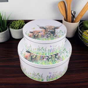 Puede incluir: Dos latas redondas para pasteles blancas con un diseño de vaca y ternero de las Tierras Altas. Las latas están apiladas una encima de la otra. La lata superior presenta una vaca de las Tierras Altas marrón y blanca con un ternero a su lado. Ambas latas tienen un diseño de hierba verde y flores alrededor de la parte inferior.