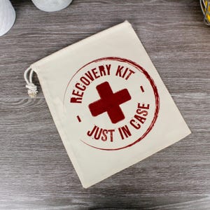 Puede incluir: Una bolsa de cordón de color crema con una cruz roja y las palabras "RECOVERY KIT JUST IN CASE" impresas en un diseño circular. La bolsa está hecha de un material similar a la lona y tiene un cierre de cordón blanco.