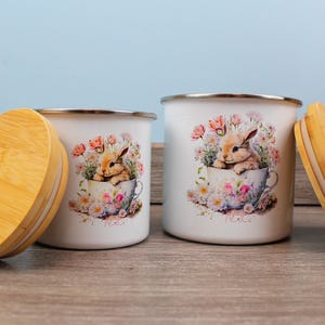 Könnte beinhalten: Zwei weiße Emaille-Dosen mit Bambusdeckeln. Die Dosen zeigen ein Blumenmuster mit einem Kaninchen, das in einer Teetasse sitzt. Der Text "Tea Time" ist auf den Dosen aufgedruckt.