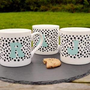 May include: Three white ceramic mugs with black polka dots and a light blue letter on each. The mugs are on a dark grey slate, with a biscuit. The letters are R, J, and another letter.