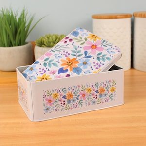 Könnte beinhalten: Eine rechteckige Aufbewahrungsbox aus weißem Metall mit floralem Design. Der Deckel und die Vorderseite der Box zeigen bunte Blumen und grüne Blätter. Die Box ist geöffnet und zeigt ein schwarzes Innere.