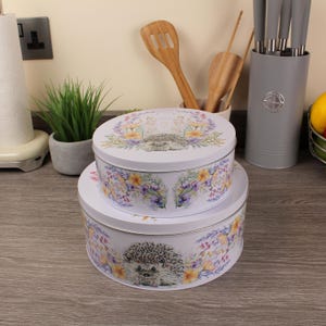 May include: Two white, round metal storage tins with lids, stacked. Each tin has a floral design with yellow, purple, and green accents, and a hedgehog illustration. The tins are on a wooden surface.