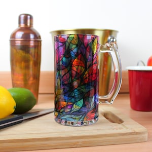 Puede incluir: Un jarro de cerveza de vidrio colorido con un diseño de vitral. El jarro tiene un asa y está hecho de vidrio transparente. El diseño presenta una variedad de colores, incluyendo rojo, naranja, amarillo, verde, azul y morado.