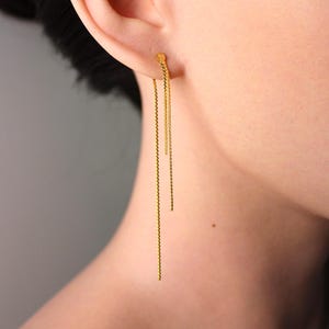 Puede incluir: Pendientes de cadena dorados. Los pendientes presentan un pequeño perno que se fija a la oreja, con tres delicadas cadenas colgando. Las cadenas son finas y cuelgan, creando un aspecto simple pero elegante.