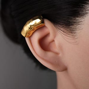 Könnte beinhalten: Ein goldfarbener Ohrklemmen-Schmuck mit gehämmerten Textur. Der Ohrschmuck wird am Ohr getragen und hat eine gebogene Form. Der Hintergrund ist einfarbig grau. Der Ohrschmuck ist ein Schmuckstück.