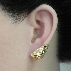 May include: A gold-colored earring with a textured surface is worn on an ear. The earring has a unique, irregular shape and a shiny finish. The earring is a stud style, designed to sit close to the earlobe.