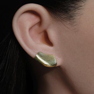 Könnte beinhalten: Ein goldfarbener Ohrschmuck mit strukturierter Oberfläche. Der Ohrschmuck hat eine geschwungene, organische Form und wird am Ohr getragen. Das Schmuckstück ist ein modernes Accessoire.