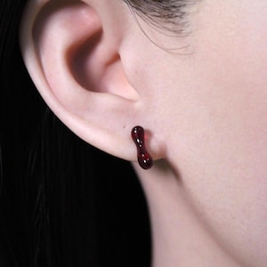 Puede incluir: Primer plano de una oreja con un pendiente alargado de color rojo oscuro. El pendiente tiene un acabado liso y brillante y una forma única y orgánica. El pendiente es de un color rojo intenso y rico.