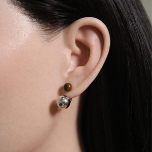 May include: A pair of earrings, one with a small, round, brown stone and the other with a large, shiny, silver ball. The earrings are worn on an ear, with the silver ball hanging below the earlobe.
