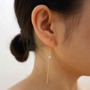 May include: A gold chain earring with a small diamond at the end. The earring is hanging from the earlobe of a person.