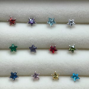 May include: Twelve star-shaped nose rings with different coloured gemstones. The rings are arranged in three rows of four. The colours of the gemstones are red, purple, blue, green, light blue, dark blue, yellow, and clear.