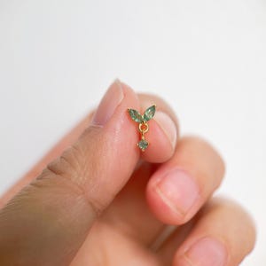 May include: A delicate gold earring featuring a leaf-like design with two green gemstones and a dangling, smaller green gemstone. The earring is held between fingers against a white background, showcasing its intricate details and elegant style.
