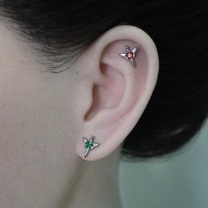 Op de afbeelding: Zilverkleurige oor piercings in de vorm van een zwaard met vleugels. Eén piercing heeft een rode edelsteen en de andere heeft een groene edelsteen. De piercings zitten op een oor.