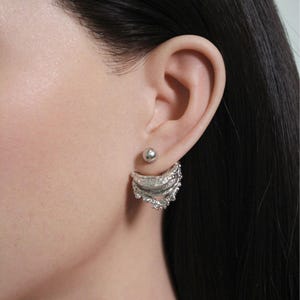 Puede incluir: Un par de pendientes plateados. Uno es un pequeño perno de bola, y el otro es un pendiente más grande, texturizado, en forma de media luna. Los pendientes se llevan en la oreja de una persona, con el pendiente más grande colocado detrás del lóbulo.