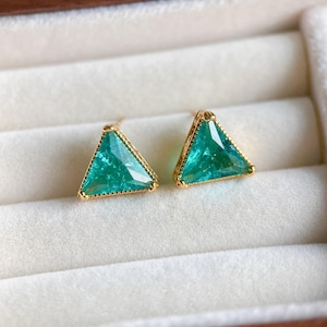 May include: A pair of gold-colored triangle-shaped earrings with a vibrant teal-green gemstone. The earrings are set in a gold frame with a textured border. The earrings are displayed in a white jewelry box.