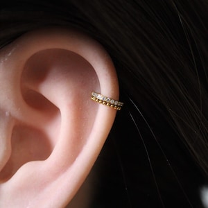 Op de afbeelding: Een gouden oormanchet met twee rijen. De bovenste rij heeft kleine, heldere edelstenen, terwijl de onderste rij een kralenontwerp heeft. De manchet wordt gedragen op het bovenste kraakbeen van het oor, met het oor en donker haar op de achtergrond.