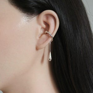 May include: A silver ear cuff with a dangling teardrop-shaped charm. The ear cuff wraps around the upper ear, connected to the charm by a delicate chain. The jewelry is shown against a background of dark hair and skin.