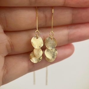 May include: Gold-colored dangle earrings. Each earring features three textured, circular charms suspended from a delicate chain. The earrings have a polished finish, reflecting light. The design is simple yet elegant, suitable for various occasions.