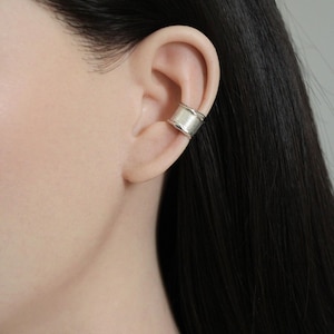 May include: A silver ear cuff with a textured, ridged design. The cuff wraps around the ear cartilage, creating a modern and minimalist look. The jewellery is shown on a person's ear, highlighting its shape and style.