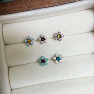 May include: A collection of six flower-shaped stud earrings. Each earring features a central gemstone surrounded by petals in various colors, including blue, purple, and green. The earrings are displayed in a white jewelry box.